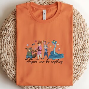 K&ouml;nnte beinhalten: Orangefarbenes T-Shirt mit einer Cartoon-Illustration von Tierfiguren. Das Design umfasst einen Biber, einen Fuchs, ein Kaninchen und eine Schlange, mit dem Text "Anyone can be anything" in einer Schriftart.
