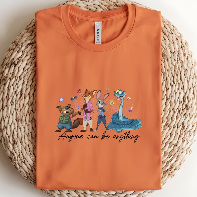 K&ouml;nnte beinhalten: Orangefarbenes T-Shirt mit einer Cartoon-Illustration von Tierfiguren. Das Design umfasst einen Biber, einen Fuchs, ein Kaninchen und eine Schlange, mit dem Text "Anyone can be anything" in einer Schriftart.