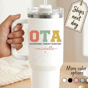 May include: White insulated tumbler with a handle, a clear lid, and a straw. The tumbler features the letters "OTA" in coral, mint green, and gold, with the words "Occupational Therapy Assistant" below. The name "michelle" is written in a cursive font.