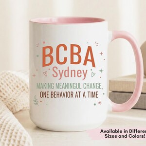 May include: White ceramic mug with a pink interior and handle. The mug features the text "BCBA Sydney" in orange and pink, along with the phrase "MAKING MEANINGFUL CHANGE, ONE BEHAVIOR AT A TIME." Decorative elements include hearts and stars.