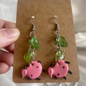 May include: A pair of handmade dangle earrings. Each earring features a pink pig charm with black accents, dangling from a silver hook. Green leaf-shaped beads and clear beads add a touch of whimsy. The earrings are displayed on a brown card.