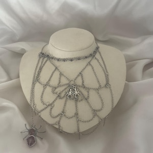 May include: A silver-toned necklace with a spiderweb design. The necklace features a spider pendant and a dangling spider with a purple gemstone body. The necklace is displayed on a mannequin against a white background.