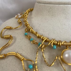 May include: A gold necklace with detailed wirework and small turquoise beads. The necklace has a large, pearlescent pendant and multiple strands. The design is intricate and layered, creating a unique and elegant accessory. The necklace is a statement piece.