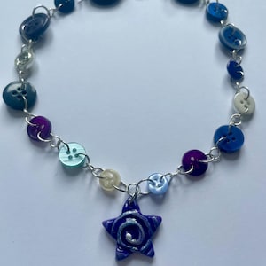 May include: A necklace constructed from a silver-toned metal chain featuring various circular buttons in shades of blue, purple, and white. A single star-shaped pendant with a blue and silver swirl pattern hangs from the center of the chain.