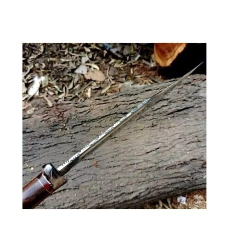 K&ouml;nnte beinhalten: Nahaufnahme einer silbernen Messerklinge mit einem Holzgriff. Die Klinge hat ein strukturiertes Muster und liegt auf einem St&uuml;ck Holz.