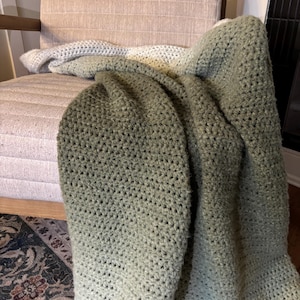 May include: A hand-crocheted blanket in shades of green and white is draped over a beige upholstered chair. The blanket's texture is visible, with a gradient effect from dark green to light green and white.