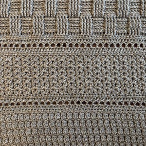 May include: A detailed view of a grey crocheted blanket, showcasing diverse stitch techniques. The design features basketweave sections, openwork rows, and dense star-shaped patterns. The blanket's texture appears soft and inviting, suitable for home use.