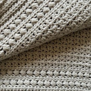 May include: A close-up of a crocheted blanket with a textured pattern. The blanket is a light gray color and has a soft, fuzzy texture.