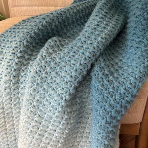 May include: A handmade, textured crochet blanket in shades of blue, draped over a beige chair. The blanket's color transitions from light blue to a deeper teal, showcasing a cozy, handcrafted design.