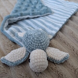 Può includere: Una morbida coperta di sicurezza e un giocattolo per bambini nei toni del blu e del bianco. La coperta presenta un motivo a righe e un angolo strutturato e increspato. Il giocattolo attaccato è una creatura rotonda e sfocata con quattro ali e una piccola testa.