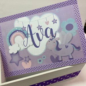 May include: A white wooden box with a purple lid featuring the name "Ava" in script. The lid is decorated with a unicorn, rainbow, stars, and clouds. Purple polka dot ribbon trims the lid and box.