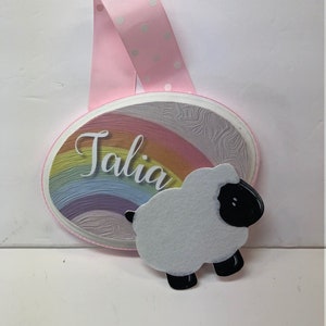 May include: A pink and white polka dot ribbon hangs from a round wooden sign with a rainbow design. The sign says "Talia" and has a white felt sheep with black eyes and hooves.