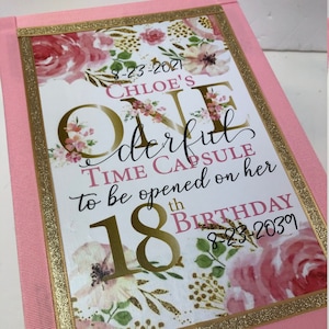 May include: A pink time capsule box with gold lettering and floral accents. The text reads "Chloe's One Wonderful Time Capsule to be opened on her 18th Birthday." The box is decorated with pink ribbon and gold glitter trim.