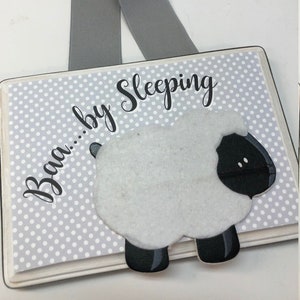 May include: A white wooden sign with gray polka dots and a black and white felt sheep. The sign says "Baa... by Sleeping" and has a gray ribbon for hanging.
