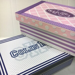 May include: Two decorative storage boxes with lids. The top box is pink with white polka dots and a purple and white striped pattern. The bottom box is gray with a chevron pattern and blue and white stripes. The top box has the text "Harper Lilley" on it. The bottom box has the text "Colin Lee" on it.