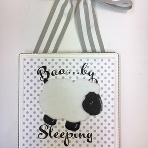 May include: A white wooden sign with a gray and white polka dot background. The sign says "Boo...by Sleeping" with a black and white sheep illustration. The sign is hanging from a gray and white striped ribbon.