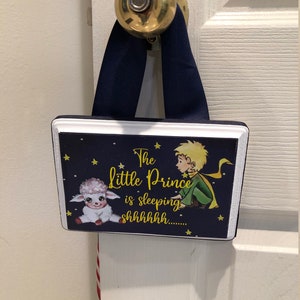 May include: A blue and white wooden sign with a picture of the Little Prince and his sheep. The sign reads "The Little Prince is sleeping shhhhhhh..."