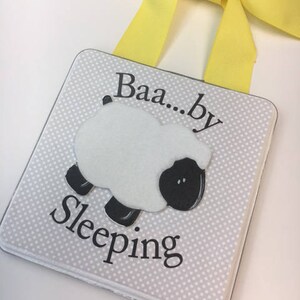 May include: A white square sign with a black and white sheep illustration and the text "Baa...by Sleeping". The sign has a yellow ribbon attached to the top for hanging.