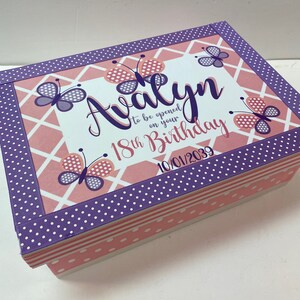 May include: A purple and pink keepsake box with a butterfly design. The box has the text "To be opened on your 18th Birthday 10/01/2039" and the name "Alayn" written in cursive.
