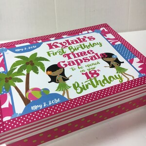May include: A pink and white wooden box with a colorful design featuring two hula girls, palm trees, and a beach ball. The box has a label that reads "Kylak's First Birthday Time Capsule to be opened on your 18th Birthday" with the dates "May 2, 2018" and "May 2, 2036".