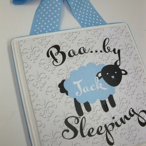 May include: A white wooden sign with a blue and white polka dot ribbon hanging from the top. The sign has a black and white sheep with a blue body and the text "Baa...by Jack Sleeping" on it.