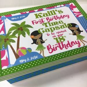 May include: A white box with a green, pink, and blue patterned design. The box is decorated with a green ribbon and has the text "Kaili's First Birthday Time Capsule to be opened on your 18th Birthday" and the dates "May 2, 2018" and "May 2, 2036".