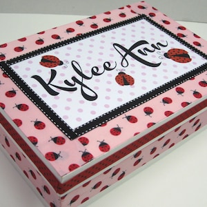 May include: A pink and white wooden box with a black and white ladybug pattern. The lid has a black frame with a white background and pink polka dots. The name "Kylee Ann" is written in black cursive lettering.