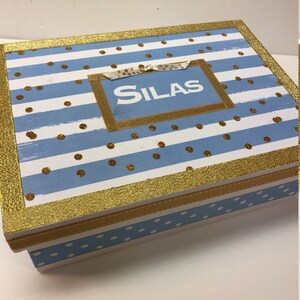 May include: A blue and white striped wooden box with gold glitter accents. The box has a gold ribbon and a label with the name "SILAS" on it.