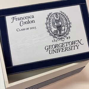 May include: White wooden box with a blue ribbon and white polka dots around the lid. The lid has a white background with black text that reads "Francesca Conlon Class of 2025" and a black and white seal with the text "1789 Georgetown University".