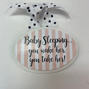 May include: A white oval sign with pink and white stripes and the text "Baby Sleeping... you wake her, you take her!" hanging from a white ribbon with black polka dots.