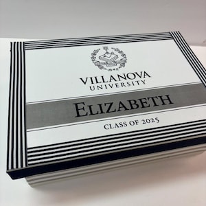 May include: A white box with black and white stripes around the edges. The box has a silver label with the text "Villanova University" and "Elizabeth" in black text. The label also has the text "Class of 2025" in black text. The box has a crest with the text "Veritas Unitas Caritas" and "1842" in black text.