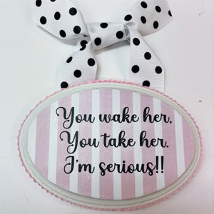 May include: Pink and white striped oval wooden sign with the text "You wake her, You take her. I'm serious!!" hanging from a white ribbon with black polka dots.
