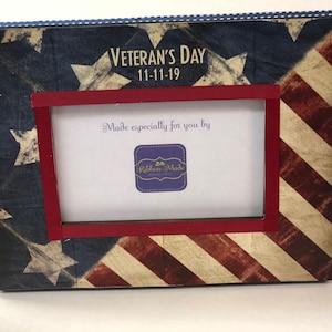 May include: A wooden picture frame with a red, white, and blue American flag design. The frame has a red ribbon border and the text "Veteran's Day 11-11-19" is printed on the top. The text "Made especially for you by Ribbon-Made" is printed on the bottom.