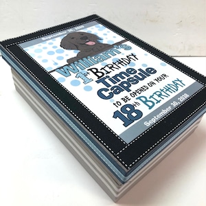 May include: A gray and blue striped box with a black border and a white label. The label says "William's 1st Birthday Time Capsule to be opened on your 18th Birthday September 30, 2038".
