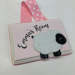 May include: Pink wooden door sign with white polka dots and the text "Emma's Room" in black cursive font. A white sheep with black legs and face is attached to the sign.