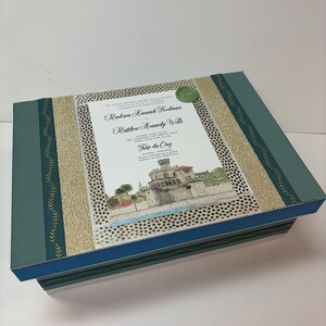May include: A green and gold wedding invitation box with a white card inside. The card has a wax seal and the text "Madison Hannah Freedman and Matthew Kennedy Wills" and "Forte da Cruz".