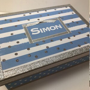 May include: A blue and white striped wooden box with silver glitter accents and white polka dots. The box has a silver glitter label with the name "SIMON" written in white.