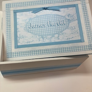 May include: A white wooden box with a blue and white checkered ribbon and a blue ribbon with a knot. The lid has a blue and white checkered background with a white oval with the text "James Harold" in blue cursive writing.