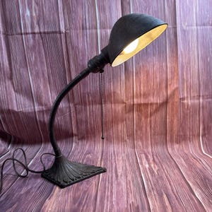 Puede incluir: Una lámpara de escritorio negra antigua con cuello flexible y base decorativa. La lámpara tiene una pantalla curva y una cadena. La bombilla está encendida, proyectando un brillo cálido. La lámpara está sobre una superficie de madera.