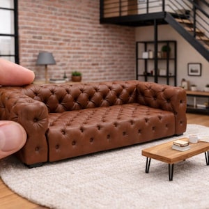 May include: Miniature brown leather sofa with button tufting, a small wooden coffee table, and a white rug. The sofa is being held by a hand. The scene is set in a room with a brick wall and a black staircase.