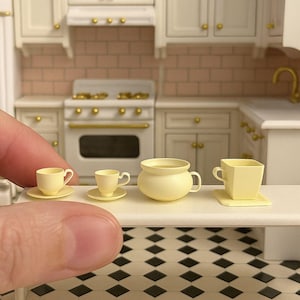 May include: Miniature yellow teacups and a small pot are arranged on a white surface in a dollhouse kitchen setting. The kitchen features a white stove, cabinets, and a pink brick backsplash. The floor has a black and white checkered pattern.