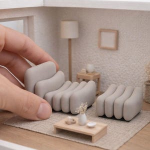 Può includere: Scena di soggiorno in miniatura con un divano e un pouf grigio chiaro. La scena comprende un tavolino, una lampada e un tappeto. L'arredamento è all'interno di una casa delle bambole. Una mano tiene il divano.
