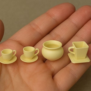 May include: A collection of miniature, pale yellow teacups, saucers, and a small pot are arranged in a hand. The set includes two traditional teacups with saucers, a small pot, and a square teacup with a matching square saucer.