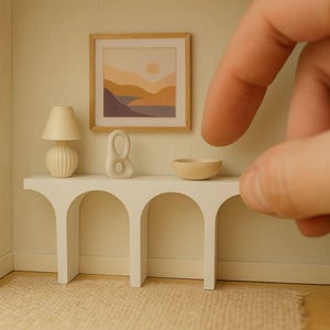 May include: Miniature interior scene featuring a white console table with arched supports, a lamp, a sculpture, and a small bowl. A framed landscape painting hangs above the table. The scene is set in a small room with a neutral color palette.