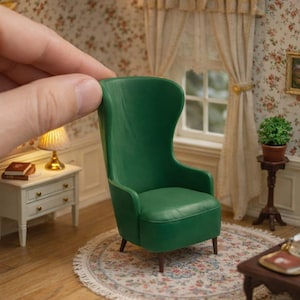 May include: Miniature green wingback chair. The chair is the focal point of a dollhouse living room scene. Other items include a small side table, a lamp, a rug, and a small table with a tea set.