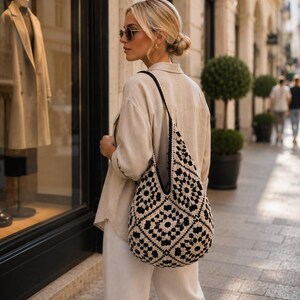 Pode incluir: Uma bolsa hobo de croch&ecirc; preta e creme com um padr&atilde;o repetitivo de quadrados granny. A bolsa tem uma al&ccedil;a larga e um formato desestruturado e flex&iacute;vel, feita de fio com um motivo floral geom&eacute;trico distinto em cores contrastantes.