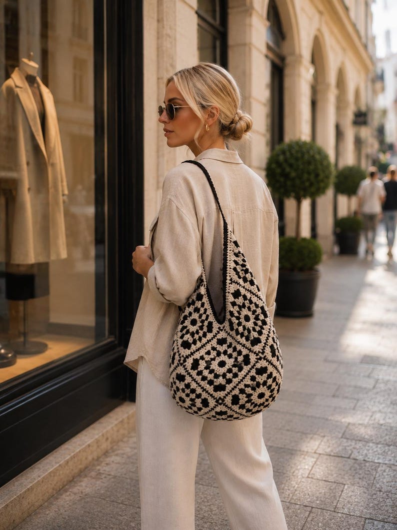 Pode incluir: Uma bolsa hobo de croch&ecirc; preta e creme com um padr&atilde;o repetitivo de quadrados granny. A bolsa tem uma al&ccedil;a larga e um formato desestruturado e flex&iacute;vel, feita de fio com um motivo floral geom&eacute;trico distinto em cores contrastantes.