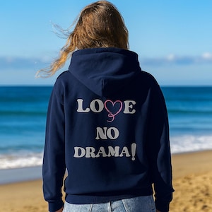 Könnte beinhalten: Ein marineblauer Kapuzenpullover mit den Worten "LOVE NO DRAMA!" in weißen Blockbuchstaben. Das Wort "LOVE" hat ein rosa Herz anstelle des "O". Der Kapuzenpullover wird an einem Strand mit dem Meer im Hintergrund getragen.