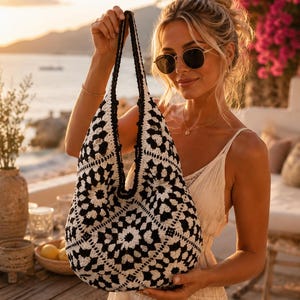 Pode incluir: Uma bolsa hobo de croch&ecirc; em preto e branco com um padr&atilde;o repetitivo de quadrados florais. A bolsa tem uma constru&ccedil;&atilde;o macia e desestruturada com uma al&ccedil;a de ombro &uacute;nica integrada e uma borda de croch&ecirc; preta s&oacute;lida na abertura superior.