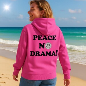 Könnte beinhalten: Ein leuchtend pinkfarbener Kapuzenpullover mit dem schwarzen Aufdruck "PEACE NO DRAMA!". Das "O" in "NO" ist ein Smiley. Der Pullover wird an einem Strand getragen, mit Meer und Himmel im Hintergrund.